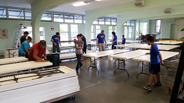 kids and adults wipe down and stack plastic folding tables in a large hall