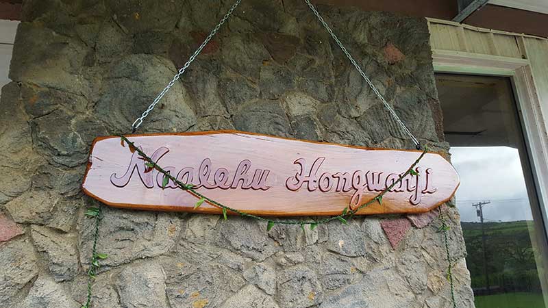 Naalehu Hongwanji wooden sign hung from a chain with a backdrop of rock wall