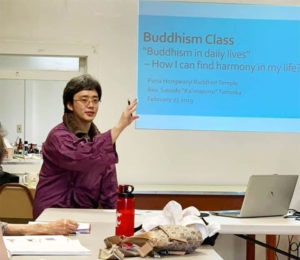 Rev. Tomioka gestures with a Buddhism class slide behind him, "Buddhism in Daily Lives"